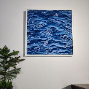 May include: A framed artwork depicting a textured, three-dimensional representation of ocean waves. The waves are rendered in shades of deep blue, with highlights suggesting sunlight. The artwork is encased in a white square frame, mounted on a white wall.