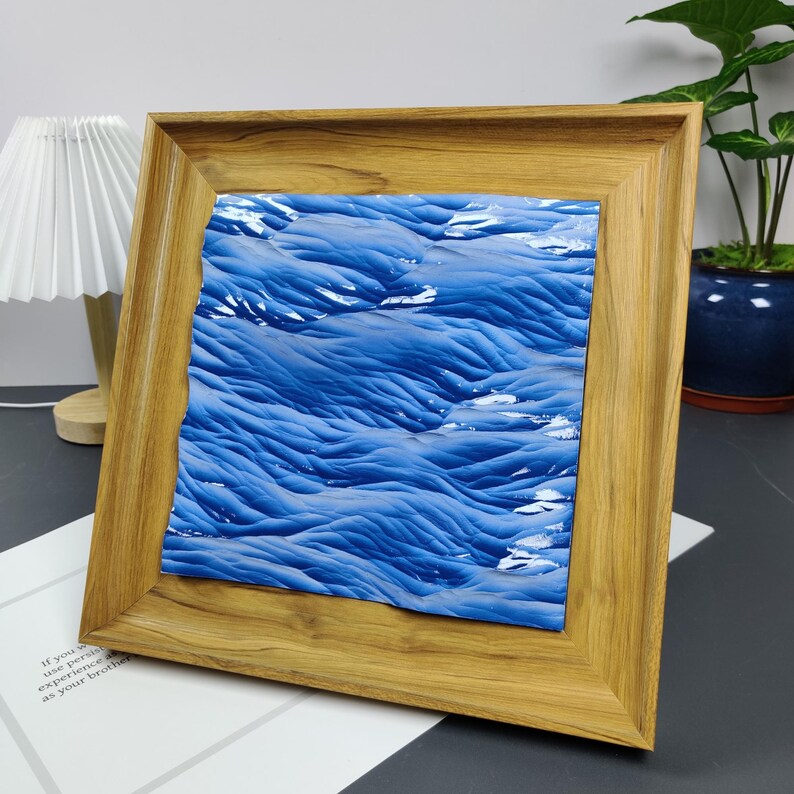 May include: A framed artwork featuring a textured blue and white abstract design. The artwork is set within a natural wood frame, creating a contrast between the cool tones of the art and the warm tones of the frame. The piece is displayed on a dark surface.