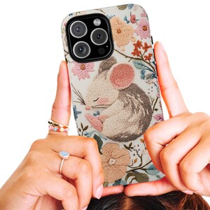 May include: A phone case with an embroidered design of a sleeping mouse. The case has a cream background with floral accents in pink, peach, and blue. The mouse is depicted in shades of grey, white, and pink.