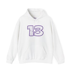 May include: A white hooded sweatshirt featuring a large number 13 printed on the chest. The number 13 has a thick purple outline and is filled with a delicate, thin-lined floral pattern in light purple, complete with a front kangaroo pocket.