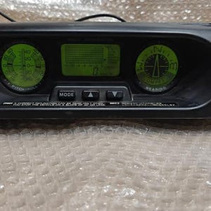 以下が含まれることがあります： 緑色に光るディスプレイを備えた黒い電子機器。左と右に2つの円形ゲージがあり、中央に長方形のディスプレイがあります。デバイスには「MODE」と矢印のラベルが付いたボタンがあります。