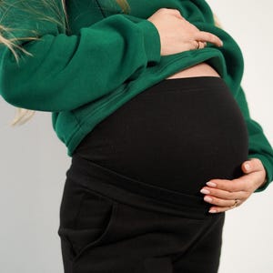 May include: A pregnant person wearing a green sweatshirt and black maternity pants. The black pants have a comfortable waistband and pockets. The sweatshirt is a solid green color.