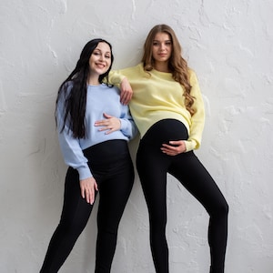 May include: Two women wearing maternity clothing. One woman wears a light blue sweater and black leggings, the other a yellow sweater and black leggings. Both are smiling and touching their bellies. They are wearing black and brown shoes.