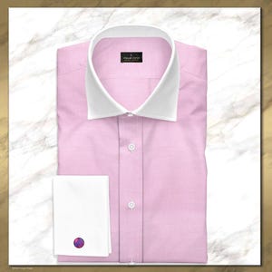 Może przedstawiać: Jasnoróżowa koszula z białym kołnierzykiem i mankietami. Koszula ma klasyczny krój z zapięciem na guziki i małą czarną metką w pobliżu kołnierzyka. Spinka do mankietu ma kolorowy, okrągły wzór.