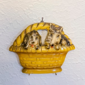 May include: A yellow ceramic wall-mounted key holder shaped like a basket with two dogs peeking out. The dogs are brown and white with black noses and brown eyes.