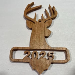 May include: A wooden deer head ornament with antlers and the year 2025 cut out below. The ornament is made of light brown wood and has a small hole at the top for hanging. The bottom of the ornament is shaped like a state.