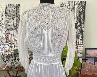 May include: A white vintage dress with intricate lace detailing. The dress features a high neckline, long sleeves, and a tiered skirt. The lace patterns are visible on the bodice, sleeves, and skirt, creating a delicate and elegant look. The dress is displayed on a mannequin.