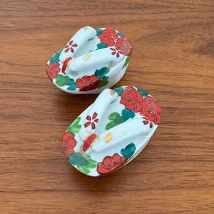 May include: A pair of white ceramic geta sandals with red and green floral designs. The sandals are traditional Japanese footwear.
