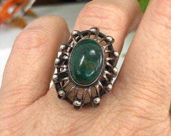 May include: Silver-toned ring featuring an oval, dark green stone. The ring has a decorative, openwork design with small, round accents around the stone. The ring is worn on a finger.