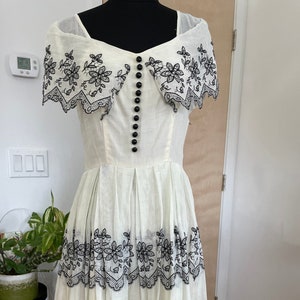 May include: Cream-colored vintage dress with black floral embroidery. The dress features a square neckline, a row of black buttons, and a tiered skirt. The floral embroidery is along the neckline and skirt hem.
