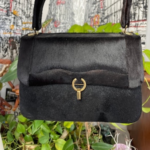 May include: Black handbag with a curved handle and a gold-colored clasp. The bag has a rectangular shape with a flap closure and a textured surface. The handle is attached with gold-colored hardware.