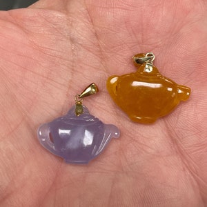 May include: Two jade teapot pendants with gold accents. The pendant on the left is a light purple color, and the pendant on the right is a bright orange color.