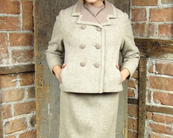 May include: A beige tweed skirt suit with a double-breasted jacket and matching knee-length skirt. The jacket has large, round buttons and a contrasting collar. The outfit is paired with black heels.