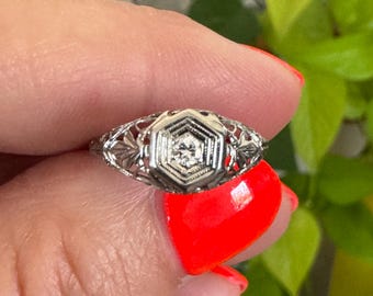 May include: A delicate silver ring with a hexagonal setting featuring a small, clear diamond. The ring has intricate filigree details and a vintage style.