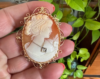 Könnte beinhalten: Ovale Kamee-Brosche mit dem Profil einer Frau, geschnitzt in Creme- und Orangetönen. Der goldfarbene Rahmen hat ein verziertes Filigran-Design. Ein kleiner Diamantanhänger hängt an der Kamee. Ein klassisches Schmuckstück.