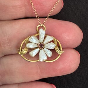 May include: A gold-toned pendant necklace featuring a flower-shaped brooch with white, irregular petal-like elements surrounding a small, round, reddish-brown center stone. The brooch is set within a gold-toned oval frame with leaf-like accents.