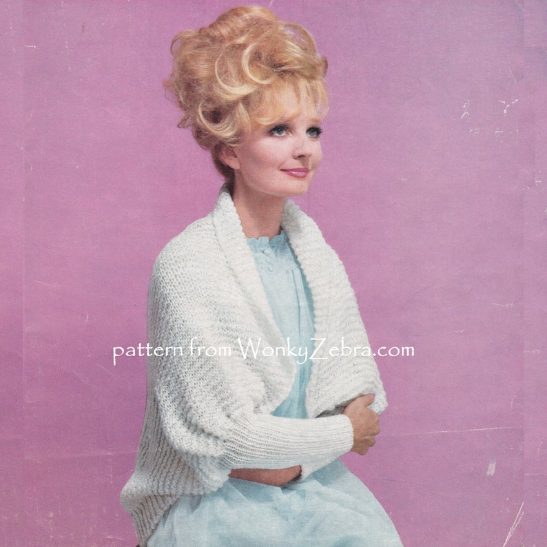 May include: A white knitted cardigan with a wide, open collar and long sleeves. The cardigan is worn by a woman with blonde hair styled in a bouffant. The background is a solid pink.