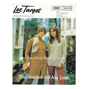 May include: Two women wearing crocheted garments. The woman on the left is wearing a long, mustard yellow coat with a white crocheted scarf. The woman on the right is wearing a shorter, cream-colored cardigan. Both garments feature a crocheted Aran pattern. The text "The Crochet ARAN Look" is printed at the bottom of the image.