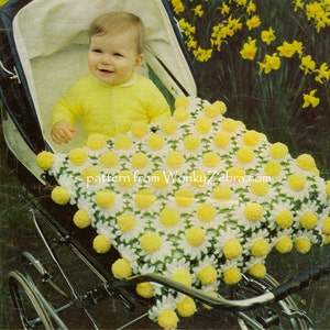 May include: A baby in a stroller is covered with a white and yellow daisy-patterned blanket. The blanket has yellow pom-poms and is surrounded by a black stroller. The baby is wearing a yellow outfit. The text "pattern from WonkyZebra.com" is visible.