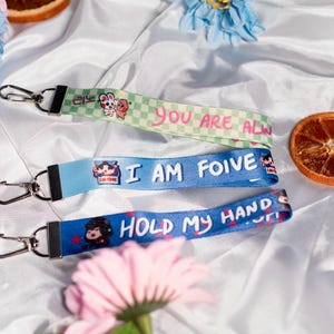 Könnte beinhalten: Drei farbige Schlüsselbänder mit Metallverschlüssen. Die Schlüsselbänder sind grün, blau und dunkelblau, jedes mit einer anderen Grafik und Text: "YOU ARE ALW", "I AM FOIVE" und "HOLD MY HANDS". Die Schlüsselbänder liegen auf einer weißen Satin-Oberfläche mit Blumen und getrockneten Orangenscheiben.