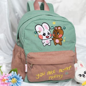 May include: A sage green and dusty rose corduroy backpack featuring an embroidered white rabbit and brown bear character design on the front. The front pocket displays the yellow embroidered text, "YOU ARE ALWAYS PRETTY," with a small star accent.