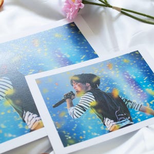 May include: Two concert photo prints featuring a performer singing into a microphone. The performer is wearing a striped long-sleeve shirt and a black vest. The background is a blue and yellow stage with confetti. A pink and blue flower and dried orange slices are also in the image.