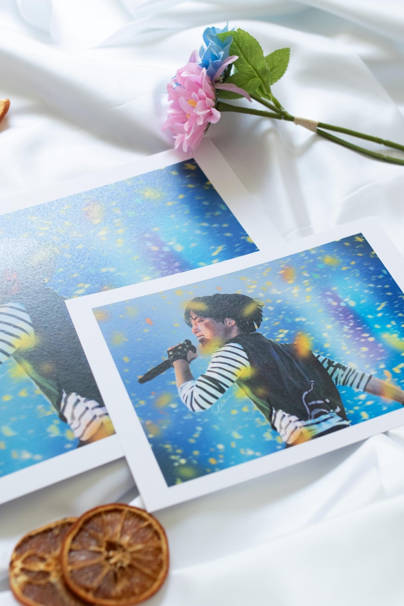 May include: Two concert photo prints featuring a performer singing into a microphone. The performer is wearing a striped long-sleeve shirt and a black vest. The background is a blue and yellow stage with confetti. A pink and blue flower and dried orange slices are also in the image.
