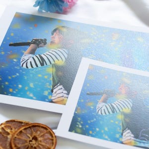 May include: Two prints featuring a person singing into a microphone, wearing a striped shirt. The prints are on a white surface, with dried orange slices and a blue and pink flower arrangement. The background is a soft, white fabric.