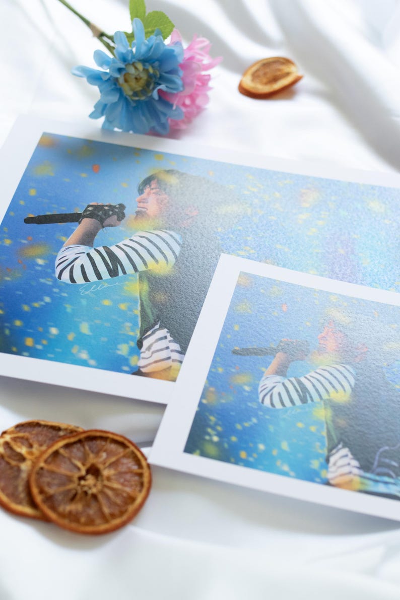 May include: Two prints featuring a person singing into a microphone, wearing a striped shirt. The prints are on a white surface, with dried orange slices and a blue and pink flower arrangement. The background is a soft, white fabric.