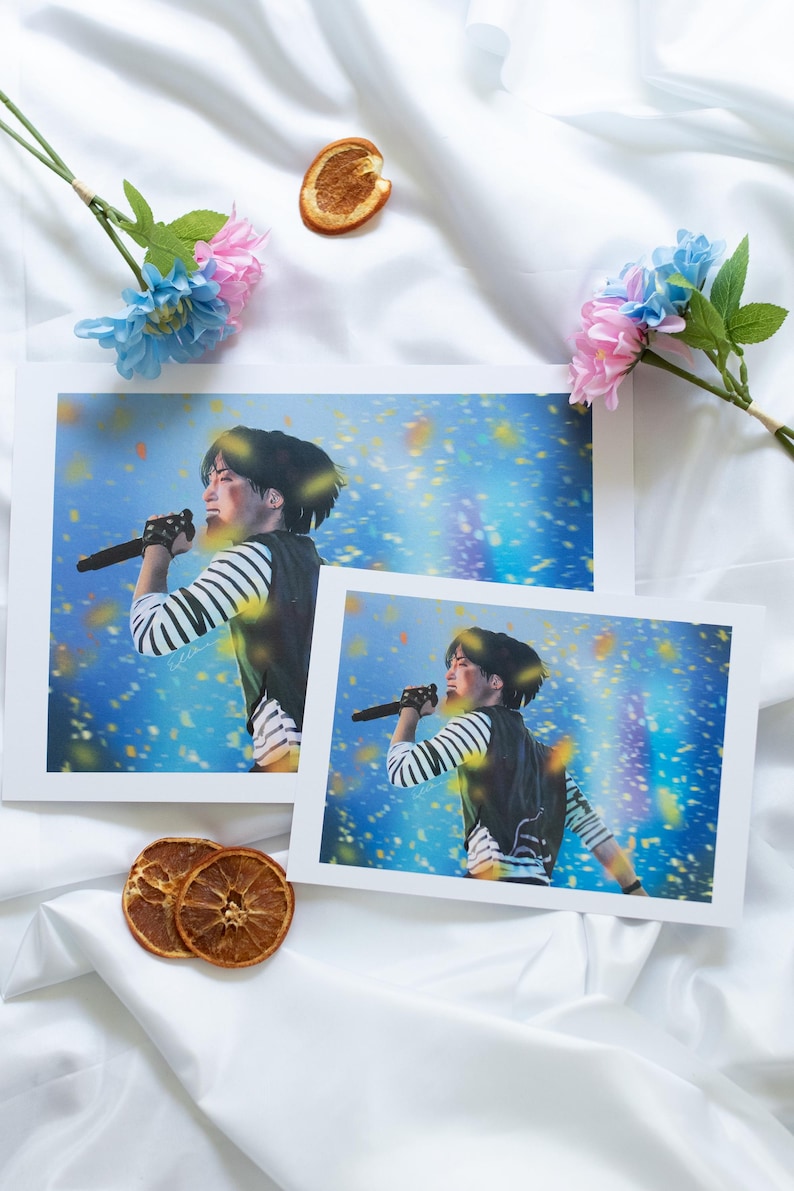 May include: Two prints of a person singing into a microphone, wearing a striped shirt and vest, against a blue background with confetti. The prints are on a white surface with flowers and dried orange slices.