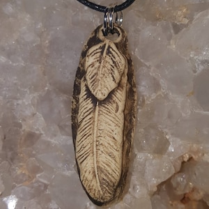 May include: A pendant featuring a carved feather design on a light tan, textured surface with dark brown accents. The pendant is oval-shaped and hangs from a black cord by a silver-toned metal ring. The feather motif is embossed on the front.