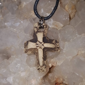 May include: A textured, off-white and dark brown cross pendant with a weathered appearance, featuring a central star-like engraving. The cross is suspended from a black cord by a silver-toned metal jump ring, creating a simple necklace design.