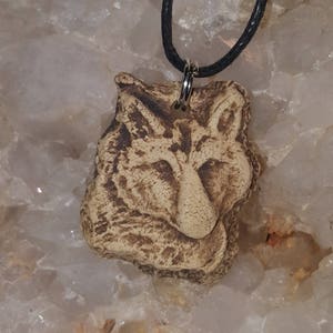 May include: A tan, textured clay pendant shaped like a fox head with dark brown detailing in the recessed areas. The pendant is suspended from a thin, black cord by a silver-toned metal jump ring, creating a rustic and earthy appearance.