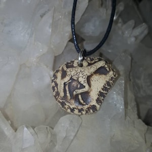 May include: A round, beige ceramic pendant featuring a dark brown embossed giraffe figure. The pendant is suspended from a thin black cord and has a textured, uneven edge with dark brown detailing around the perimeter of the circular piece.