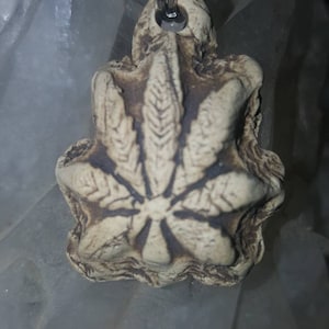 May include: A beige, textured pendant with a carved leaf design featuring seven distinct leaflets radiating from a central point. The pendant has a dark, weathered finish in the recessed areas and is suspended from a thin, black cord by a metal ring.