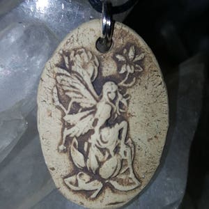 May include: An oval-shaped beige pendant featuring a dark brown embossed design of a winged figure sitting on a flower. The pendant has a hole at the top with a silver-toned metal jump ring attached to a thin black cord necklace.