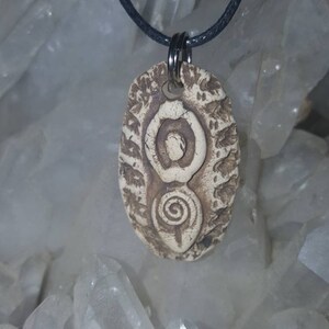 May include: An oval-shaped clay pendant with a textured, beige surface featuring a carved figure-eight design with a spiral at the bottom. The pendant is suspended from a thin black cord by a silver-toned metal jump ring and split ring.