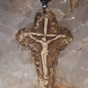 May include: A tan and brown textured clay crucifix pendant featuring a raised figure of Jesus on the cross. The pendant has a rough, uneven surface and is suspended from a simple black cord by a silver-toned metal jump ring and bail.
