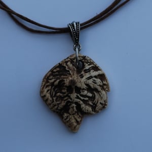 May include: A pendant necklace featuring a textured, cream-colored, organic-shaped piece with dark brown recessed detailing. The pendant is suspended from a silver-toned metal bail on a thin, dark brown cord that loops around the top.