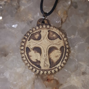 May include: A circular pendant featuring a raised cross design with dark brown recessed areas and a light beige textured surface. The edge of the pendant has a beaded border detail. It is suspended from a thin black cord through a metal jump ring.
