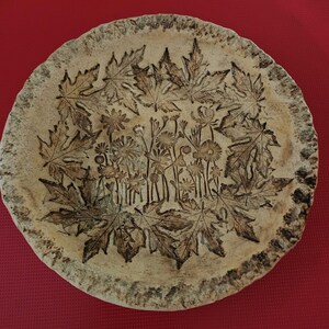 May include: A round, beige ceramic plate featuring a textured, uneven rim and a dark brown embossed design. The center displays a cluster of various wildflowers, while the outer border is decorated with a ring of detailed maple leaf impressions.