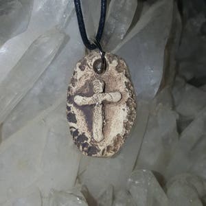 May include: An oval-shaped pendant with a textured, beige, and brown surface featuring a raised cross design in the center. The pendant is suspended from a thin, black cord and is positioned against a background of clear, jagged quartz crystals.