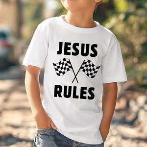 Pode incluir: Camiseta branca com o texto "JESUS RULES" em letras pretas e duas bandeiras quadriculadas cruzadas. A camiseta é usada por uma criança, com as mãos nos bolsos, contra um fundo desfocado ao ar livre.