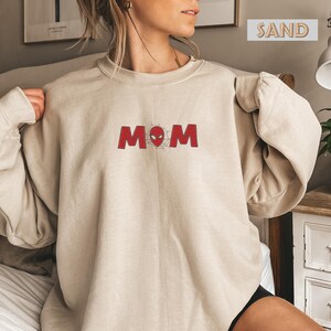 Pode incluir: Camisola cor de areia com a palavra "MOM" em vermelho, com um desenho de teia de aranha no lugar do "O". A camisola &eacute; de gola redonda e tem mangas compridas. A palavra "SAND" est&aacute; impressa numa caixa no canto superior direito.
