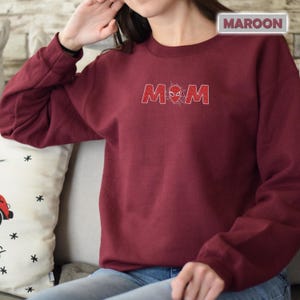 Pode incluir: Camisola bord&ocirc; com a palavra "MOM" em fonte glitter vermelha. A letra "O" &eacute; substitu&iacute;da por um desenho de teia de aranha. A palavra "MAROON" est&aacute; em um ret&acirc;ngulo branco no canto superior direito.