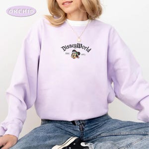 May include: A lilac sweatshirt featuring the words "DisneyWorld" and a Mickey Mouse graphic. The sweatshirt has a crew neck and long sleeves. The text "EST. 1971" is below the DisneyWorld logo. The brand name "ORCHID" is in the upper left corner.