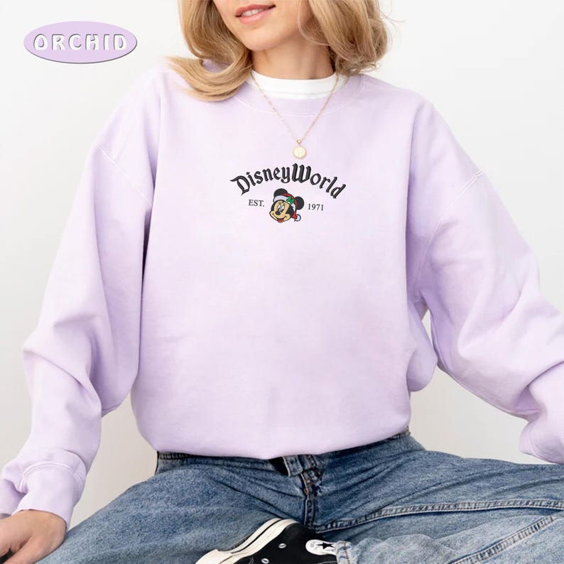 May include: A lilac sweatshirt featuring the words "DisneyWorld" and a Mickey Mouse graphic. The sweatshirt has a crew neck and long sleeves. The text "EST. 1971" is below the DisneyWorld logo. The brand name "ORCHID" is in the upper left corner.