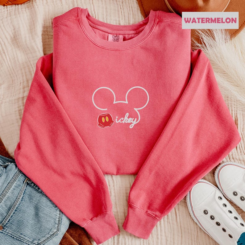 Peut inclure: Un sweat-shirt rose corail avec un contour blanc de la t&ecirc;te de Mickey Mouse et le mot "Mickey" en &eacute;criture cursive. Le sweat-shirt a des manches longues et un col rond. Le mot "WATERMELON" est imprim&eacute; sur une &eacute;tiquette rectangulaire.