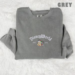 May include: A grey sweatshirt featuring white embroidered text reading "DisneyWorld", "EST. 1971", and a Mickey Mouse graphic in a Santa hat. The word "GREY" is printed in the upper right corner.