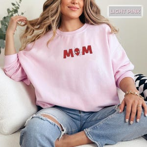 Pode incluir: Camisola rosa claro com a palavra "MOM" em vermelho, com um desenho de teia de aranha. A camisola &eacute; de cor lisa e tem mangas compridas. O texto "LIGHT PINK" est&aacute; num ret&acirc;ngulo no canto superior direito.